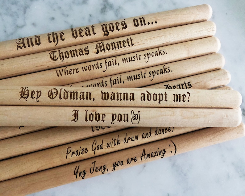 Peut inclure: Un ensemble de baguettes de batterie en bois avec des messages personnalis&eacute;s grav&eacute;s dessus. Les messages incluent "And the beat goes on...", "Thomas Monnett", "Where words fail, music speaks.", "Hey Oldman, wanna adopt me?", "I love you", "Praise God with drum and dance!" et "Ung Jeng, you are Amazing :)".
