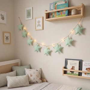 May include: A child's bedroom with a wooden bed, cushions, and a star-shaped garland. The wall has framed pictures and wooden shelves with books. The star garland is made of light green fabric.