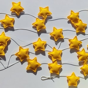 Puede incluir: Una guirnalda decorativa de estrellas amarillas de fieltro ensartadas en un hilo blanco. Las estrellas están dispuestas en un patrón ondulado. Las estrellas son de color amarillo brillante y tienen cinco puntas. El fondo es una superficie blanca lisa.