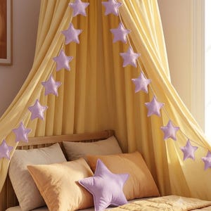 May include: A light purple star-shaped garland hangs over a yellow fabric canopy draped above a bed. The bed features two beige pillows, one orange pillow, and a light purple star-shaped throw pillow resting on a yellow quilted bedspread.