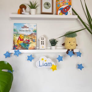 May include: A white wall-mounted shelf displays a children's room decor. Items include a Winnie the Pooh book, a coloring book, a framed picture, a mushroom plush, a pineapple plush, and a cloud-shaped banner with the name "Liam" and star-shaped ornaments.