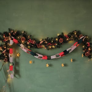 May include: A Christmas garland with fairy lights, pine cones, and red berries. A paper chain in red, black, and white gingham patterns hangs below. Gold star decorations are scattered on the green wall.