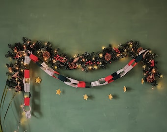 May include: A festive Christmas garland with a string of lights, pinecones, and red berries. A paper chain in red, black, and white gingham patterns hangs below. Gold star ornaments are scattered on the green wall.