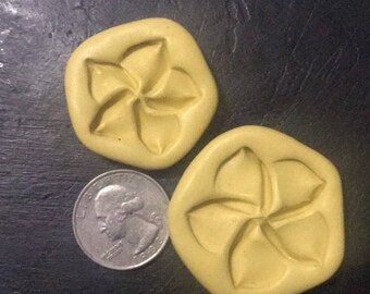 May include: Two yellow, flower-shaped molds with a quarter coin for size comparison. The molds have a five-petal design and are set against a dark background. The coin shows a profile of a person.