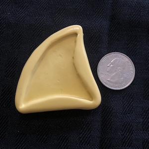May include: A yellow silicone mold shaped like a shark fin. The mold is about the size of a quarter.