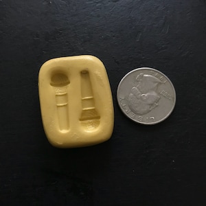 May include: A yellow silicone mold with two cavities shaped like microphones. The mold is about the size of a US quarter.