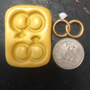 May include: A yellow silicone mold with two impressions of interlocking rings with a diamond in the center. The mold is next to a quarter and a pair of gold interlocking rings with a diamond.