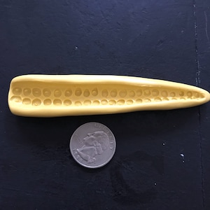 May include: A yellow silicone mold shaped like an ear of corn with a row of indentations for making corn on the cob.