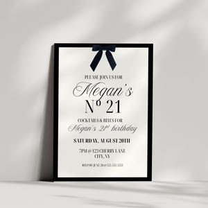 May include: A framed invitation for Megan's 21st birthday. The invitation has a black frame, with a navy blue bow at the top. The text is in an elegant script, with the words "Megan's N&ordm; 21" prominently displayed. The invitation includes the date, time, and location.
