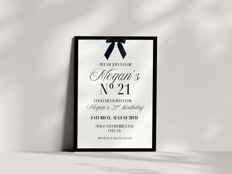 May include: A framed invitation for Megan's 21st birthday. The invitation has a black frame, with a navy blue bow at the top. The text is in an elegant script, with the words "Megan's N&ordm; 21" prominently displayed. The invitation includes the date, time, and location.