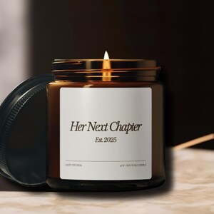 May include: A brown glass candle jar with a white label that reads "Her Next Chapter Est. 2025". The candle is lit and the flame is visible.