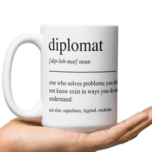 May include: White ceramic mug with a black and white definition of the word "diplomat" printed on it. The definition reads: "[dip-luh-mat] noun one who solves problems you did not know exist in ways you do not understand. see also; superhero, legend, rockstar"