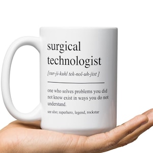 Pode incluir: Caneca de café de cerâmica branca com um design preto que diz "tecnólogo cirúrgico" e define o termo como "alguém que resolve problemas que você não sabia que existiam de maneiras que você não entende". A caneca está na mão de uma pessoa.