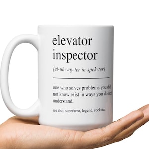 Pode incluir: Uma caneca de cerâmica branca com uma alça grande. A caneca apresenta o texto "elevator inspector" em negrito preto, juntamente com uma pronúncia fonética e uma definição. A caneca é segurada na mão de uma pessoa.