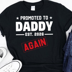 May include: Black t-shirt with white text that reads "Promoted to Daddy Est. 2026 Again". The text "Again" is in a red box.