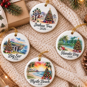 May include: Four round white ceramic Christmas ornaments featuring colorful illustrations of Joshua Tree, Big Sur, Hanalei Bay, and Myrtle Beach. Each ornament displays a decorated Christmas tree and the year 2026 with its respective location name.