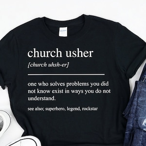 Op de afbeelding: Zwart T-shirt met witte tekst die "church usher [church uhsh-er] one who solves problems you did not know exist in ways you do not understand. see also; superhero, legend, rockstar" luidt.