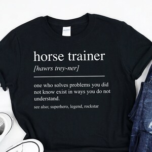 May include: Black t-shirt with white text that reads "horse trainer [hawrs trey-ner] one who solves problems you did not know exist in ways you do not understand. see also; superhero, legend, rockstar"