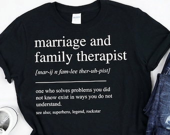 May include: Black t-shirt with white text that reads "marriage and family therapist [mar-ij n fam-lee ther-uh-pist] one who solves problems you did not know exist in ways you do not understand. see also; superhero, legend, rockstar"