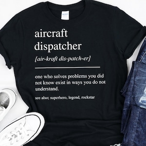 Puede incluir: Camiseta negra con el texto "aircraft dispatcher" y su definición en blanco. La camiseta se combina con vaqueros azules y zapatillas blancas. La definición dice: "alguien que resuelve problemas que no sabías que existían."