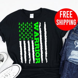 Op de afbeelding: Zwart T-shirt met een Amerikaans vlagontwerp met een groen lint en het woord "WARRIOR" verticaal gedrukt. Het shirt wordt getoond op een witte achtergrond met een paar witte sneakers en een blauwe jeans.