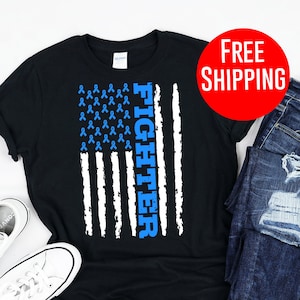 Puede incluir: Camiseta negra con un diseño de bandera americana de cinta azul y la palabra "Fighter" impresa verticalmente en el lado derecho de la bandera. La camiseta se muestra sobre un fondo blanco con un par de zapatillas blancas y unos vaqueros azules.