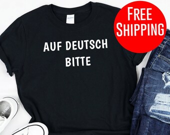 May include: Black t-shirt with white text that says "AUF DEUTSCH BITTE". The shirt is laid flat on a white surface with a pair of white sneakers and blue jeans.