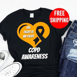 Puede incluir: Camiseta negra con un corazón y una cinta naranja. El corazón tiene el texto "His Fight Is My Fight" y el texto "COPD Awareness" está debajo del corazón.