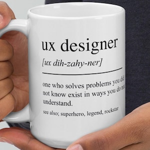 Op de afbeelding: Witte keramische mok met de tekst "ux designer" en de fonetische uitspraak ervan. De mok bevat ook een definitie van een UX-ontwerper en de woorden "superhero, legend, rockstar".