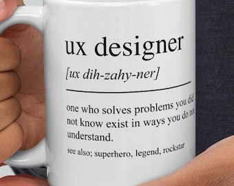 Może przedstawiać: Biały ceramiczny kubek z napisem "ux designer" i jego fonetyczną wymową. Kubek zawiera również definicję projektanta UX oraz słowa "superhero, legend, rockstar".