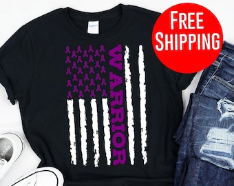 May include: Black t-shirt featuring a white and purple American flag design. The flag has purple ribbons in place of the stars and the word "WARRIOR" written vertically down the right side. The t-shirt is displayed on a white background with a pair of white trainers and blue jeans.