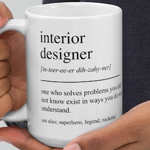 May include: White ceramic mug featuring the words "interior designer" and its phonetic spelling. The mug also includes a definition of an interior designer. The mug is held by a person.