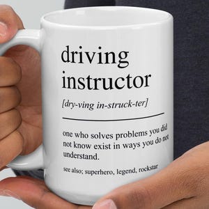Op de afbeelding: Witte keramische mok met de zwarte tekst "driving instructor". De mok bevat een fonetische uitspraak en een definitie. De onderkant van de mok vermeldt "superhero, legend, rockstar".