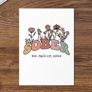 May include: A white card with the word "SOBER" in colorful, retro-style lettering, adorned with floral illustrations. Below the word is the text "Est. April 17, 2023". The card is on a wooden surface, with a keyboard and coffee cup in the background.