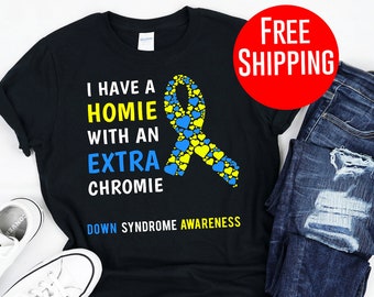 Op de afbeelding: Zwart T-shirt met de tekst "I have a homie with an extra chromie" in wit, geel en blauw. Een blauw en gele lintafbeelding staat ook op het shirt. De woorden "Down Syndrome Awareness" staan onderaan. Witte sneakers en een blauwe jeans staan ook in de afbeelding.