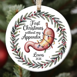 May include: A round white ceramic ornament hanging from a jute string. It features a green pine wreath with red berries, a cartoon stomach wrapped in colorful Christmas lights with a bandage, and the text First Christmas without my Appendix Lewis 2026.
