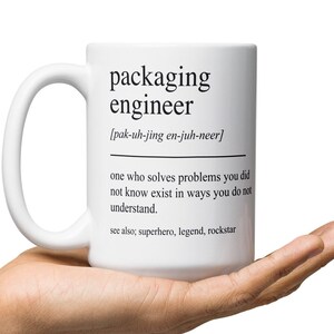 Op de afbeelding: Een witte keramische mok met een zwart ontwerp met de tekst "packaging engineer [pak-uh-jing en-juh-neer] one who solves problems you did not know exist in ways you do not understand. see also; superhero, legend, rockstar"