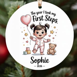 Op de afbeelding: Witte ronde kerstversiering met een cartoonillustratie van een baby die een roze hartvormige ballon vasthoudt. De tekst luidt: "The year I took my First Steps." De baby draagt een roze onesie. De naam "Sophie" en het jaar "2026" staan er ook op.