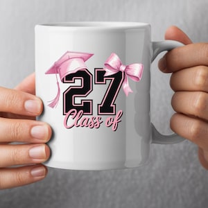 May include: White ceramic mug featuring a pink graduation cap, a pink bow, and the number "27" in black with a pink outline. The words "Class of" are written below the number in pink script. The mug is held by a person.