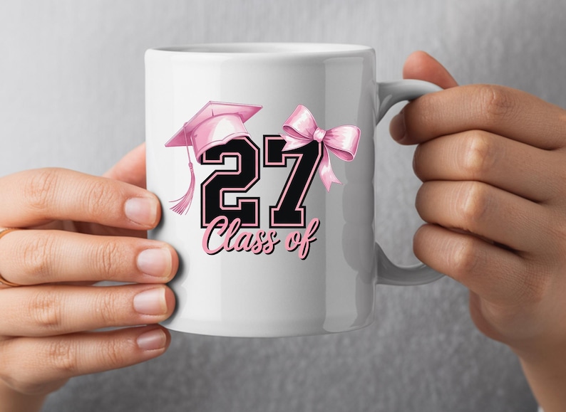 May include: White ceramic mug featuring a pink graduation cap, a pink bow, and the number "27" in black with a pink outline. The words "Class of" are written below the number in pink script. The mug is held by a person.