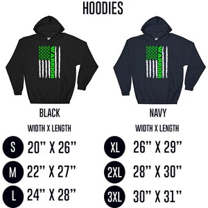 Op de afbeelding: Zwarte en marineblauwe hoodies met een wit en groen Amerikaans vlagontwerp. De vlag heeft groene lint symbolen en het woord "WARRIOR" verticaal geschreven. De hoodies zijn verkrijgbaar in de maten S, M, L, XL, 2XL en 3XL. De maattabel toont de breedte en lengte van elke maat in inches.