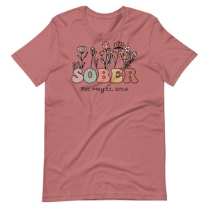 May include: A dusty rose t-shirt with the word "SOBER" in colorful bubble letters, accompanied by a floral design and the date "Est. May 11, 2016". The shirt is a short-sleeved crew neck.