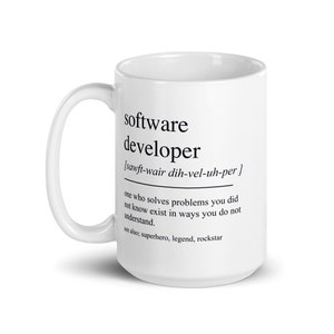 Op de afbeelding: Een witte keramische mok met een zwarte afbeelding waarop staat "Softwareontwikkelaar [sawft-wair dih-vel-uh-per] iemand die problemen oplost waarvan je niet wist dat ze bestonden, en dat op een manier die je niet begrijpt. zie ook: superheld, legende, rockster"