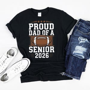 K&ouml;nnte beinhalten: Schwarzes T-Shirt mit wei&szlig;em Text, der "Proud Dad of a Senior 2026" lautet. Oberhalb des Textes befindet sich eine Grafik eines braunen und wei&szlig;en Fu&szlig;balls.