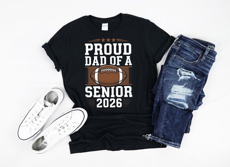 K&ouml;nnte beinhalten: Schwarzes T-Shirt mit wei&szlig;em Text, der "Proud Dad of a Senior 2026" lautet. Oberhalb des Textes befindet sich eine Grafik eines braunen und wei&szlig;en Fu&szlig;balls.