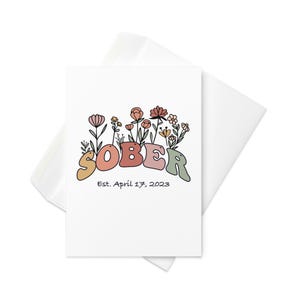 May include: A white greeting card with the word "SOBER" in colorful, retro-style lettering, adorned with floral illustrations. The card includes the text "Est. April 17, 2023" and is accompanied by a white envelope.