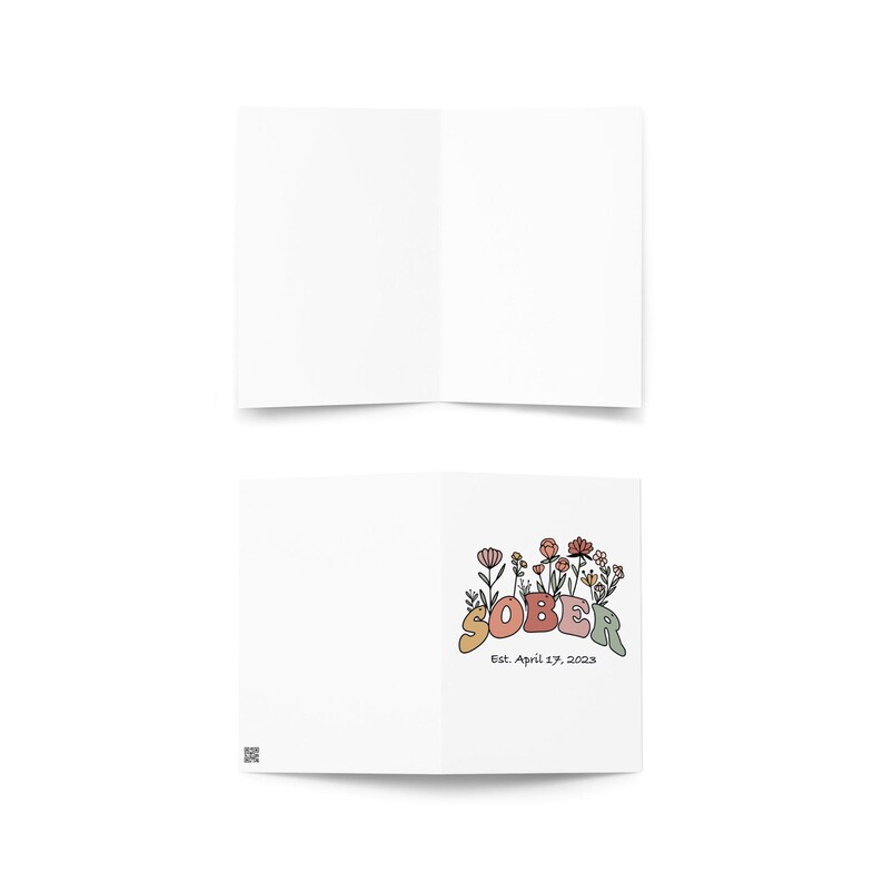 May include: A white greeting card, folded in half, with the word "SOBER" in colorful, retro-style lettering. The card is decorated with floral illustrations and the text "Est. April 27, 2023".
