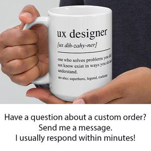 Op de afbeelding: Witte keramische mok met de tekst "ux designer" en de uitspraak ervan. De mok bevat ook een definitie van een UX-ontwerper. De mok wordt door een persoon vastgehouden.