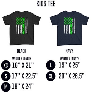 Op de afbeelding: Kinder T-shirt met groene 'Warrior'-tekst en een Amerikaans vlagontwerp. Het shirt is verkrijgbaar in zwart en marineblauw. De maten omvatten XS, S, M, L en XL. Het shirt meet 16 inch breed en 21 inch lang in maat XS, 17 inch breed en 22,5 inch lang in maat S, 18 inch breed en 24 inch lang in maat M, 19 inch breed en 25 inch lang in maat L en 20 inch breed en 26,5 inch lang in maat XL.