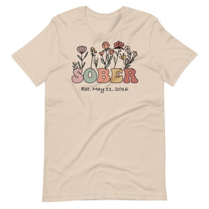 May include: A beige t-shirt with a floral design above the word "SOBER" in colorful, rounded letters. Below the word "SOBER" is the text "Est. May 11, 2016". The floral design includes pink, yellow, and orange flowers.